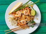 Prawn Pad Thai noodles with chopsticks. Prawn Pad Thai Noodles are a world-famous delicacy, here, this colorful traditional dish is photographed directly above on a round enameled metal white dish, with various condiments on the plate that are normally eaten together with the Pad Thai noodles. These condiments featured here are a raw banana flower, chives, bean shoots, and fresh lime. Sprinkling dried chili powder, sugar, and crushed peanuts on the dish is also very popular. This makes for a great color contrast composition with the dish set on an abstract, colorful, vibrant, worn, turquoise colored wood panel table background.
